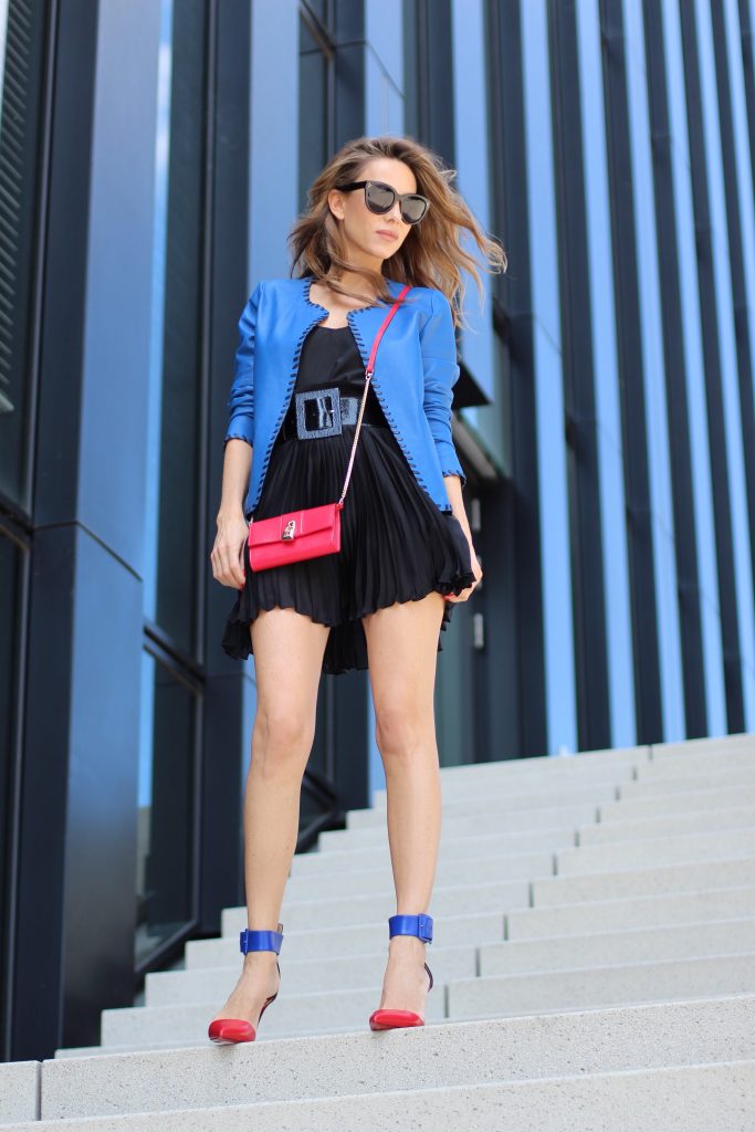 Model and Blogger Alexandra Lapp wearing a blue leather jacket from Riani, high waist pleated skirt by Givenchy, black silk top from Jadicted, a black lacquer waist belt from Dolce & Gabbana, big cat-eye 'Audrey' sunglasses from Céline, a red crossover purse bag from Patrizia Pepe and 'Harler' 100cm calf multicolor high heels from Christian Louboutin with a red sol, red front, black back and a blue little belt around the ankles, on June 2017 in Duesseldorf.
