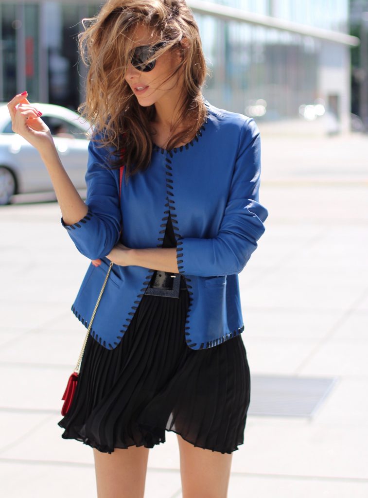 Model and Blogger Alexandra Lapp wearing a blue leather jacket from Riani, high waist pleated skirt by Givenchy, black silk top from Jadicted, a black lacquer waist belt from Dolce & Gabbana, big cat-eye 'Audrey' sunglasses from Céline, a red crossover purse bag from Patrizia Pepe and 'Harler' 100cm calf multicolor high heels from Christian Louboutin with a red sol, red front, black back and a blue little belt around the ankles, on June 2017 in Duesseldorf.
