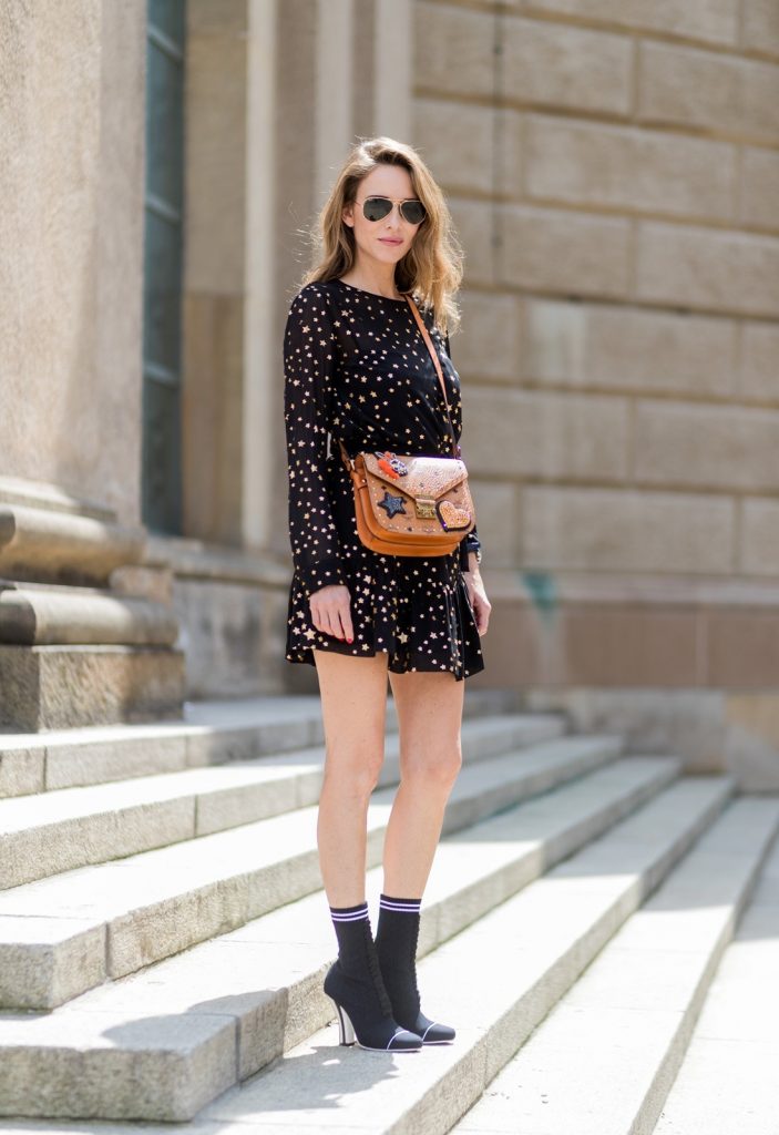 Alexandra Lapp (model, blogger) wearing Fendis SS 17 collection sock boots with a grey knit upper, with a high sculptural heel, a black silk chiffon mini dress with gilded stars from RED Valentino with a tie at the neck and a ruffle through the hem, a MCM Patricia Shoulder Bag crafted in in Visetos-print coated canvas and topped with crystalized appliqués, including the Punk Rabbit, accented with mink fur, studwork and edge painting and a laurel-engraved case latch closure and Ray Ban aviator sunglasses in gold during the Mercedes-Benz Fashion Week Berlin Spring/Summer 2018 on July 6, 2017 in Berlin, Germany.