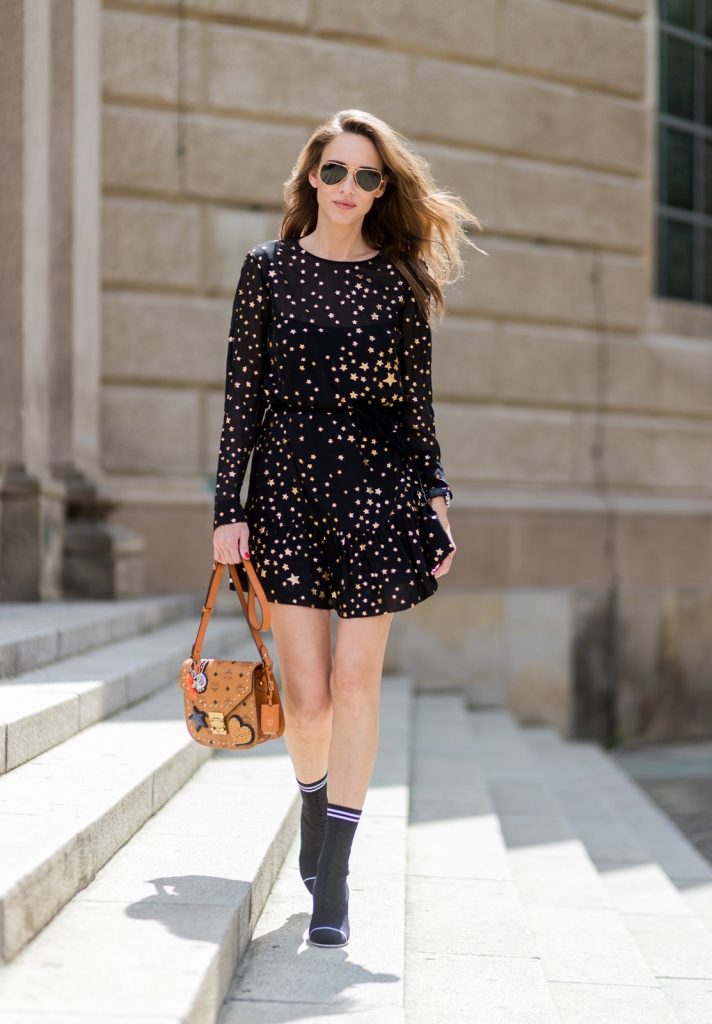 Alexandra Lapp (model, blogger) wearing Fendis SS 17 collection sock boots with a grey knit upper, with a high sculptural heel, a black silk chiffon mini dress with gilded stars from RED Valentino with a tie at the neck and a ruffle through the hem, a MCM Patricia Shoulder Bag crafted in in Visetos-print coated canvas and topped with crystalized appliqués, including the Punk Rabbit, accented with mink fur, studwork and edge painting and a laurel-engraved case latch closure and Ray Ban aviator sunglasses in gold during the Mercedes-Benz Fashion Week Berlin Spring/Summer 2018 on July 6, 2017 in Berlin, Germany.