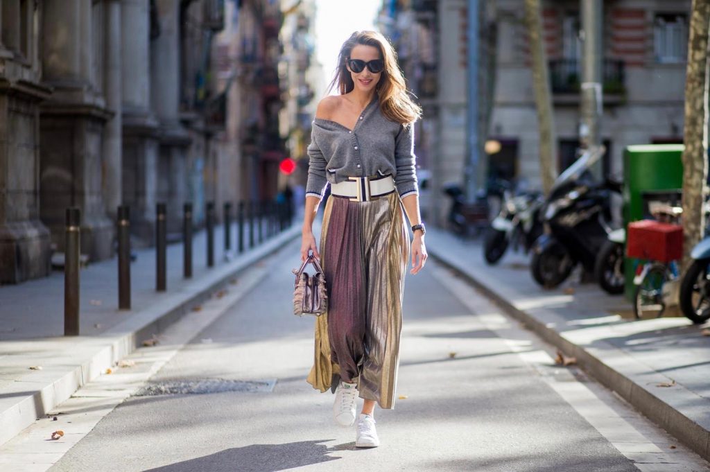 Alexandra Lapp wearing a long pleated metallic skirt with changing gold and red colors, grey cashmere cardigan with pearl buttons and blue and white rims, white and black lacquer waist belt from Balmain with gold buckle and details, Milla handbag in crystal tweed from MCM with metallic yarn and czech crystals, white and green Adidas Stan Smith sneakers and black Celine sunglasses on November 27, 2017 in Barcelona, Spain.