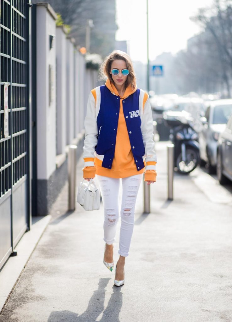 MILAN, ITALY - FEBRUARY 21: Alexandra Lapp wearing college style, a college jacket in blue, white and orange from MCM, an orange hoodie from glam-o-meter, white destroyed jeans from Frame a white Milla handbag from MCM, white pumps from Christian Louboutin and Ray-Ban sunglasses seen during Milan Fashion Week Fall/Winter 2018/19 on February 21, 2018 in Milan, Italy.