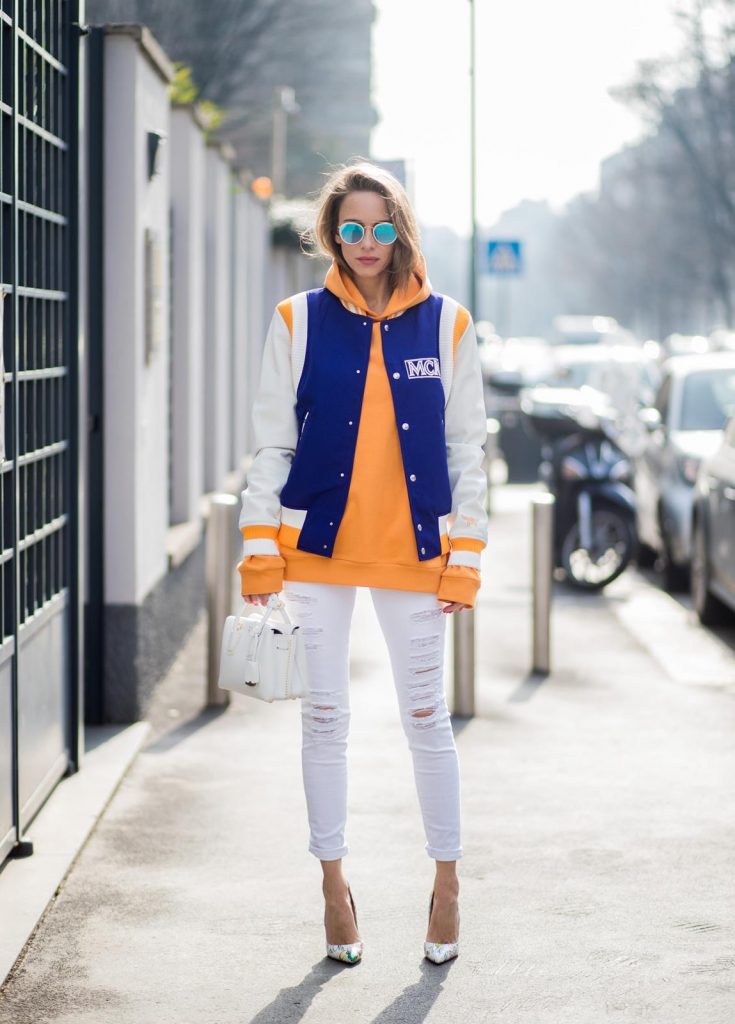 MILAN, ITALY - FEBRUARY 21: Alexandra Lapp wearing college style, a college jacket in blue, white and orange from MCM, an orange hoodie from glam-o-meter, white destroyed jeans from Frame a white Milla handbag from MCM, white pumps from Christian Louboutin and Ray-Ban sunglasses seen during Milan Fashion Week Fall/Winter 2018/19 on February 21, 2018 in Milan, Italy.
