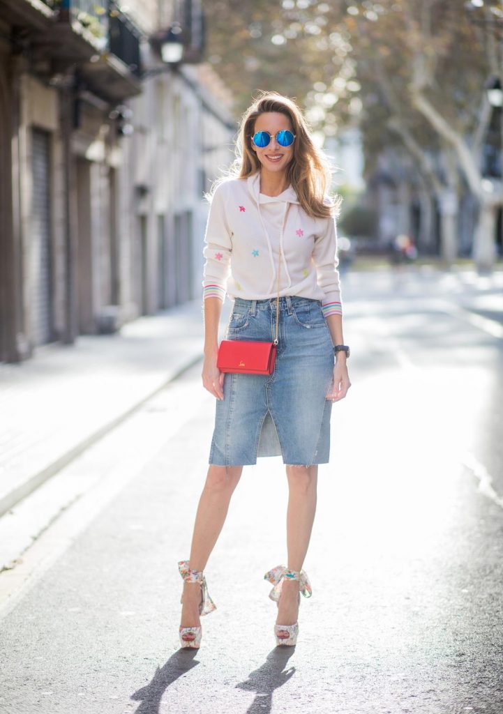 BARCELONA, SPAIN - NOVEMBER 27: Alexandra Lapp wearing a white cashmere hoodie with colored flower applications from Heartbreaker, high rise jeans skirt with slit Adriano Goldschmied, red little purse bag with gold chain from Christian Louboutin, peep-toe pump Christian Louboutin with a long satin bow, sunglasses with round blue shades from Mykita on November 27, 2017 in Barcelona, Spain. 
