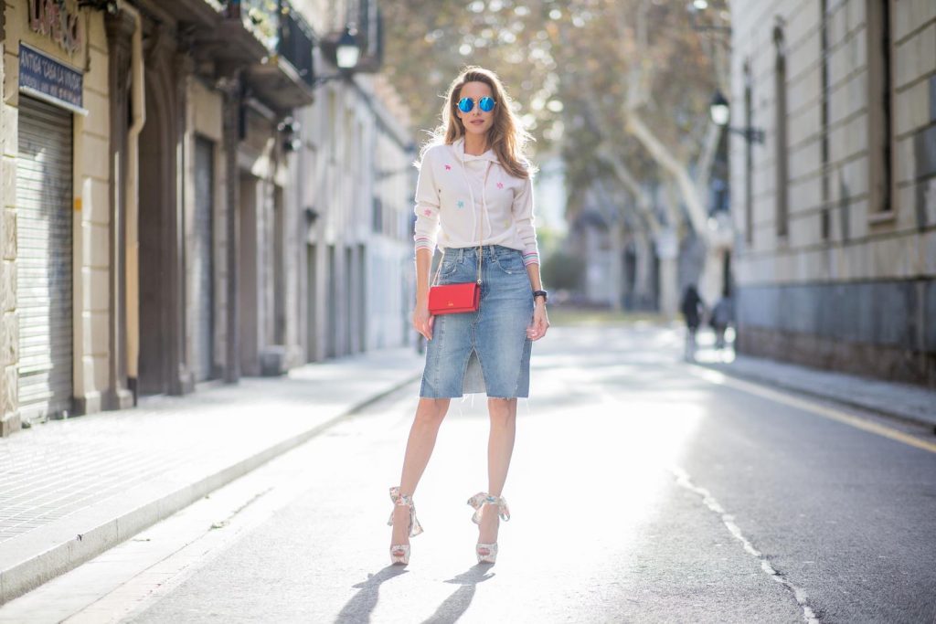 BARCELONA, SPAIN - NOVEMBER 27: Alexandra Lapp wearing a white cashmere hoodie with colored flower applications from Heartbreaker, high rise jeans skirt with slit Adriano Goldschmied, red little purse bag with gold chain from Christian Louboutin, peep-toe pump Christian Louboutin with a long satin bow, sunglasses with round blue shades from Mykita on November 27, 2017 in Barcelona, Spain. 