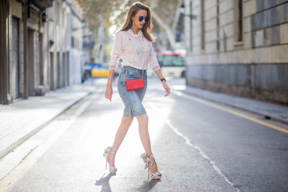 BARCELONA, SPAIN - NOVEMBER 27: Alexandra Lapp wearing a white cashmere hoodie with colored flower applications from Heartbreaker, high rise jeans skirt with slit Adriano Goldschmied, red little purse bag with gold chain from Christian Louboutin, peep-toe pump Christian Louboutin with a long satin bow, sunglasses with round blue shades from Mykita on November 27, 2017 in Barcelona, Spain.
