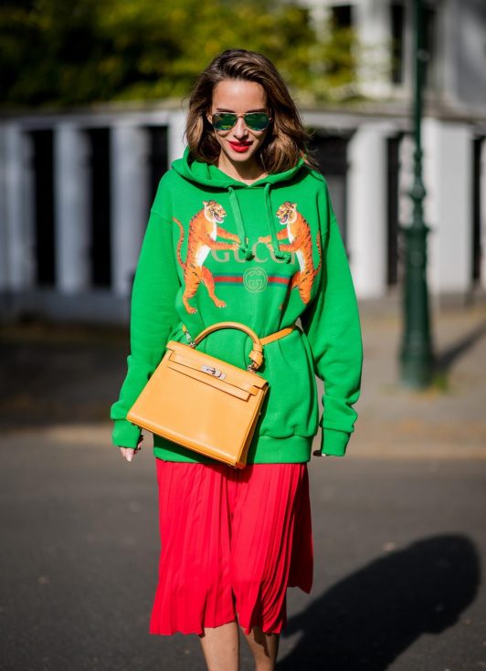 Alexandra Lapp in a Hermès Kelly Look wearing a green tiger print Gucci logo hoodie, a red pleated skirt by SET, a Hermes Birkin 30 bag in orange, Pigalle Follies pumps by Christian Louboutin and green mirrored Ray Ban sunglasses on May 3, 2018 in Duesseldorf, Germany.