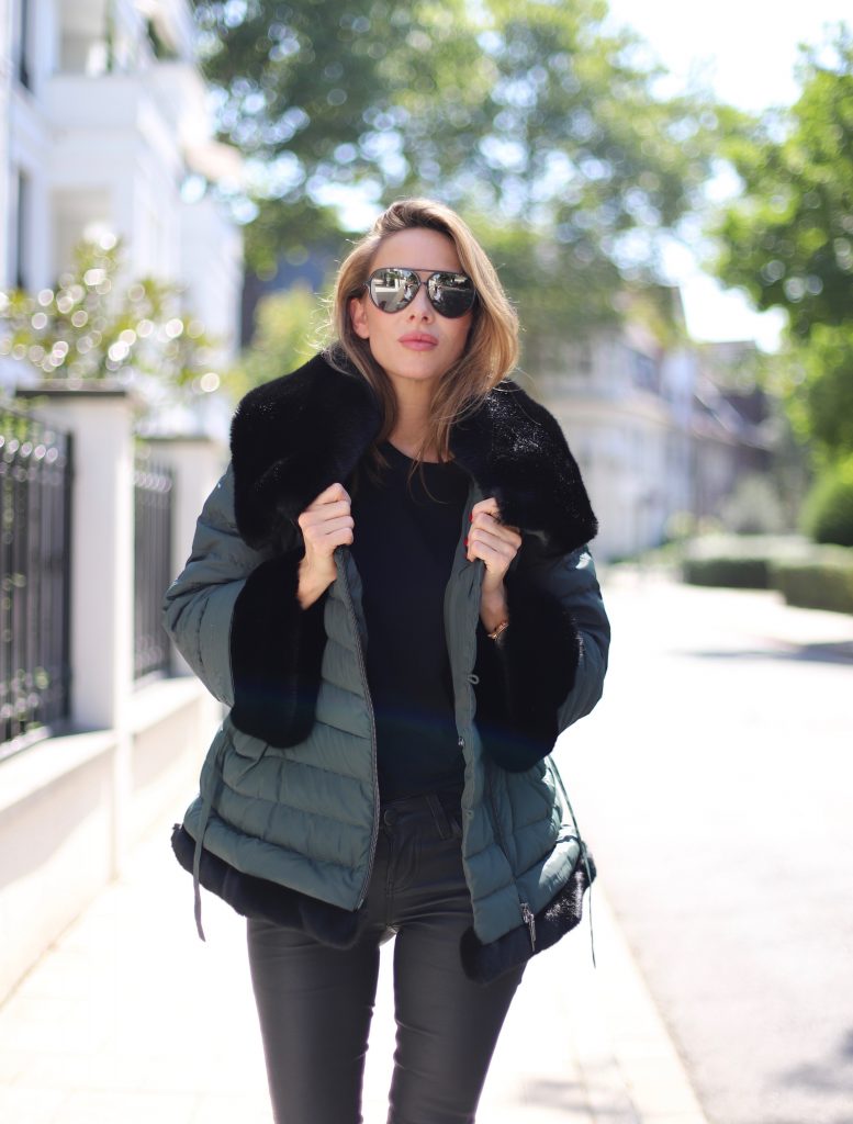 Model and Blogger Alexandra Lapp in a Faux Fur Jacket Look wearing a black down jacket with faux fur by Airfield, black leather pants by SET, black T-Shirt by James Perse, dark green leather overknee boots by Saint Laurent, black Kelly bag by Hermès and black Audrey sunglasses from Céline.