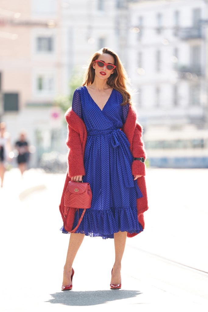 Model and blogger Alexandra Lapp with IWC Watches wearing a blue Polka-dot silk-blend dress by Valentino, Elado Cashmere and Silk-blend Cardigan in red by The Row, red Gg Marmont Quilted Leather Backpack by Gucci, red Céline Cat-Eye Sunglasses, red Gianvito Rossi heels, IWC Portugieser Chronograph and the Portofino Automatic Limited Edition by IWC.