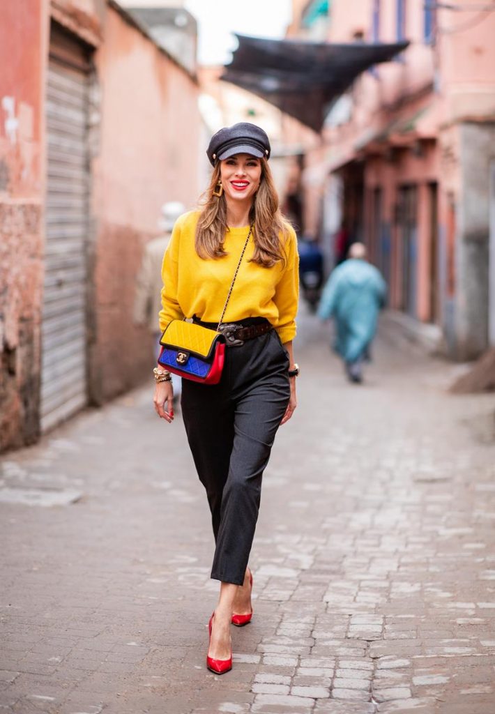 Alexandra Lapp in a Yellow Knit look, wearing a yellow crew-neck cashmere sweater from Jardin des Orangers, high-waisted trousers in dark grey by H&M, Graphic Flap Bag from Chanel in black/red/yellow/blue, brown lacquer Kieselstein Cord Crocodile belt, red lacquer pumps from Gianvito Rossi and dark grey nautical cap with buttons by ZARA on November 25, 2018 in Marrakech, Morocco.