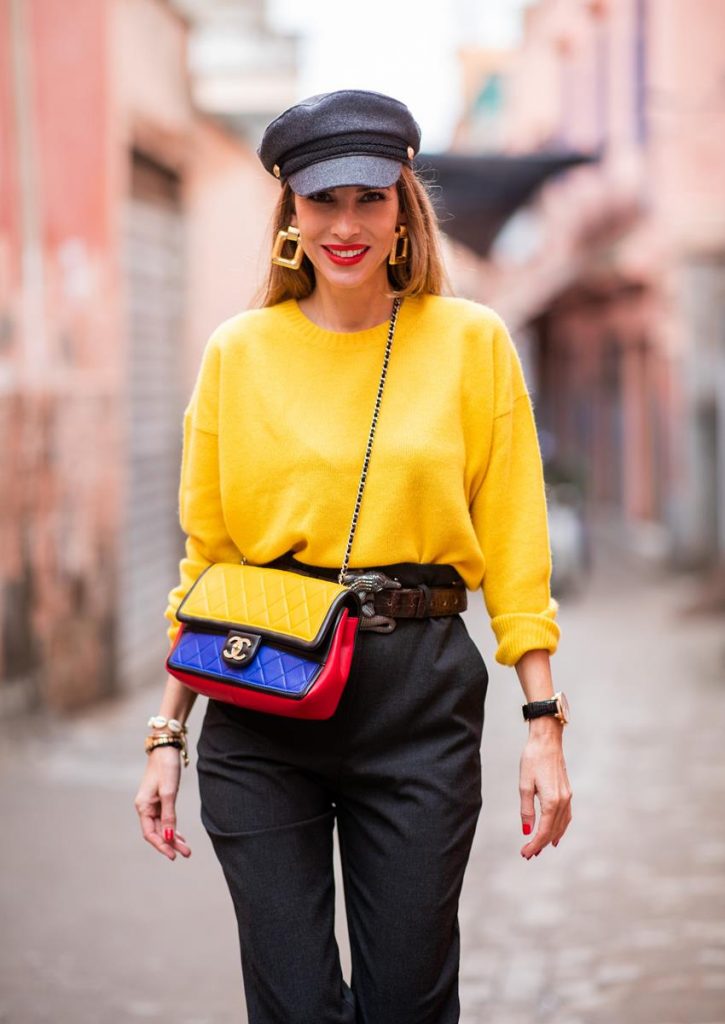  Alexandra Lapp in a Yellow Knit look, wearing a yellow crew-neck cashmere sweater from Jardin des Orangers, high-waisted trousers in dark grey by H&M, Graphic Flap Bag from Chanel in black/red/yellow/blue, brown lacquer Kieselstein Cord Crocodile belt, red lacquer pumps from Gianvito Rossi and dark grey nautical cap with buttons by ZARA on November 25, 2018 in Marrakech, Morocco.