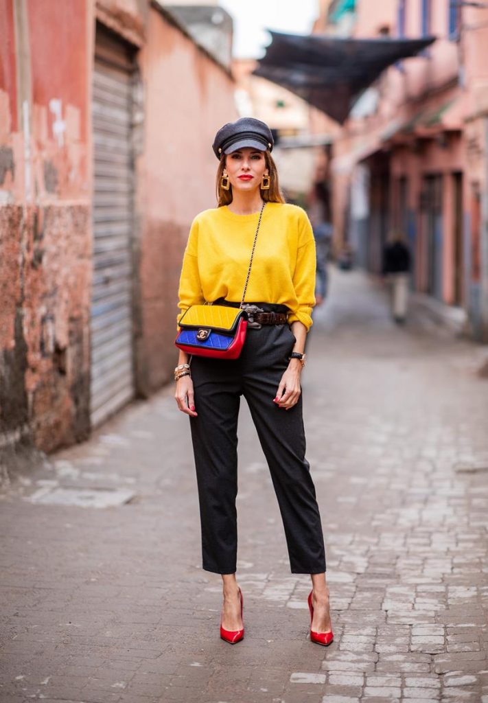  Alexandra Lapp in a Yellow Knit look, wearing a yellow crew-neck cashmere sweater from Jardin des Orangers, high-waisted trousers in dark grey by H&M, Graphic Flap Bag from Chanel in black/red/yellow/blue, brown lacquer Kieselstein Cord Crocodile belt, red lacquer pumps from Gianvito Rossi and dark grey nautical cap with buttons by ZARA on November 25, 2018 in Marrakech, Morocco.