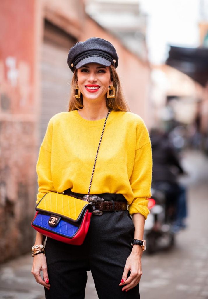 Alexandra Lapp in a Yellow Knit look, wearing a yellow crew-neck cashmere sweater from Jardin des Orangers, high-waisted trousers in dark grey by H&M, Graphic Flap Bag from Chanel in black/red/yellow/blue, brown lacquer Kieselstein Cord Crocodile belt, red lacquer pumps from Gianvito Rossi and dark grey nautical cap with buttons by ZARA on November 25, 2018 in Marrakech, Morocco.