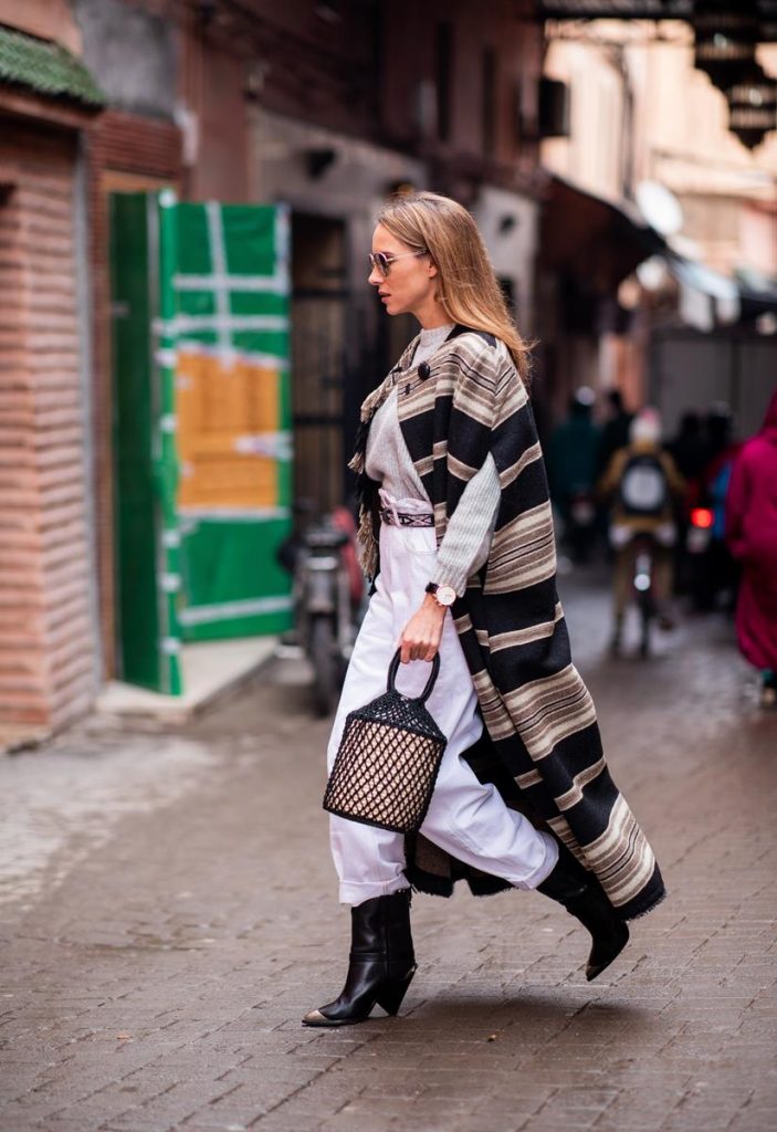Alexandra Lapp in a Poncho Style wearing a long Huan fringed striped wool cape from Isabel Marant, an oversized grey wool sweater from H&M, white high-rise boyfriend style jeans from Isabel Marant Etoile, a black leather belt with white stitches from Isabel Marant, black Cowboy boots with a silver tip from Isabel Marant, a handwoven basket bag from Sensistudio and mirrored sunglasses from Givenchy on November 25, 2018 in Marrakech, Morocco.
