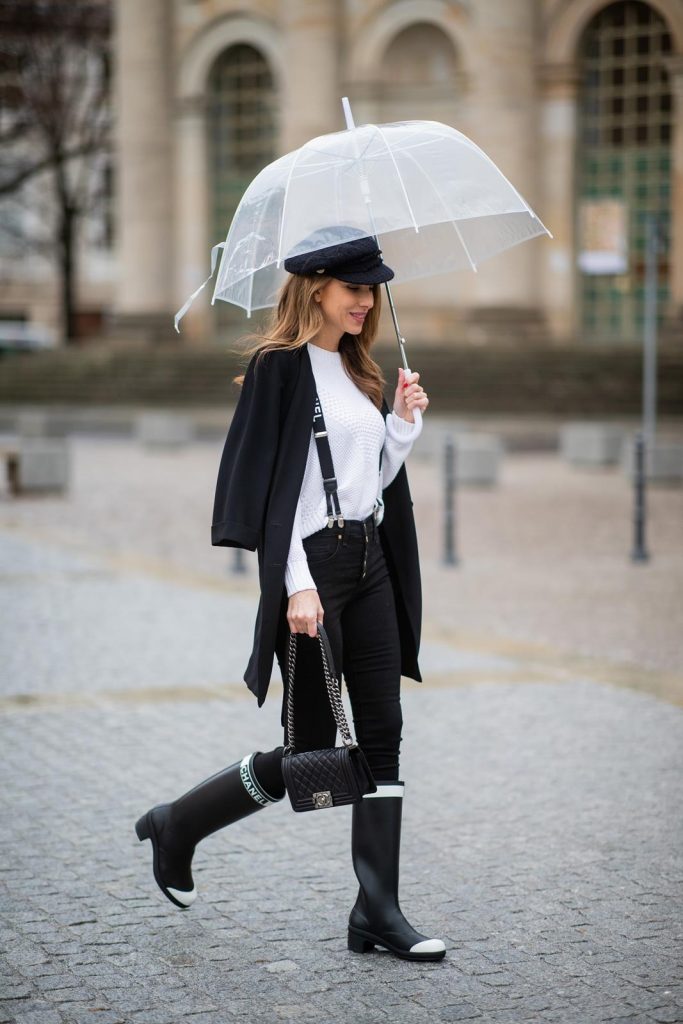 Alexandra Lapp in A Chanel Rain Boots look, wearing a soft long blazer by Steffen Schraut, over a white chunky knit sweater by H&M, black skinny pants with golden buttons by Veronica Beard, styled with Chanel logo suspenders and Chanel rain boots, the Chanel Le boy bag, a Chanel tweed baker boy cap and a transparent umbrella by Mirviory during the Berlin Fashion Week Autumn/Winter 2019 on January 15, 2019 in Berlin, Germany. (Photo by Christian Vierig/Getty Images)
