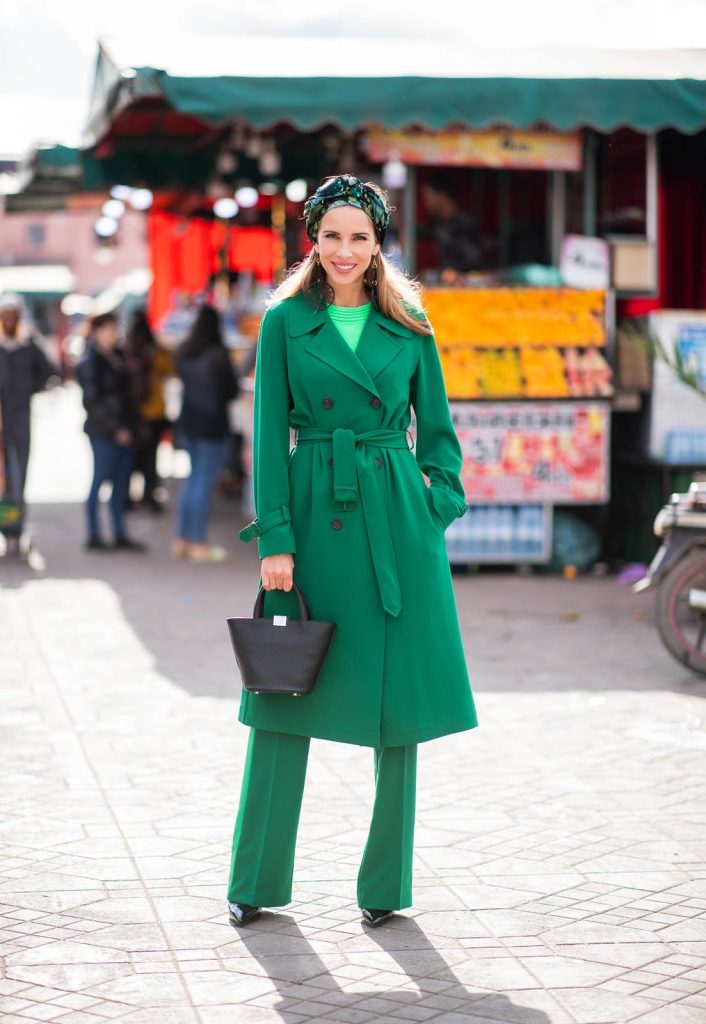 Alexandra Lapp in a Le Riad look, wearing a belted long crepe Marc Cain coat in green, a neon green sweater and crepe loose-fit pants with stripes in two green tones by Marc Cain, a black small trapeze shaped bag and a printed silk scarf in multi green shades by Marc Cain, black lacquer high heels, black sunglasses and round oversized earrings in black by Marc Cain on November 26, 2018 in Marrakech, Morocco. 