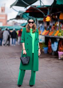 Alexandra Lapp in a Le Riad look, wearing a belted long crepe Marc Cain coat in green, a neon green sweater and crepe loose-fit pants with stripes in two green tones by Marc Cain, a black small trapeze shaped bag and a printed silk scarf in multi green shades by Marc Cain, black lacquer high heels, black sunglasses and round oversized earrings in black by Marc Cain on November 26, 2018 in Marrakech, Morocco.