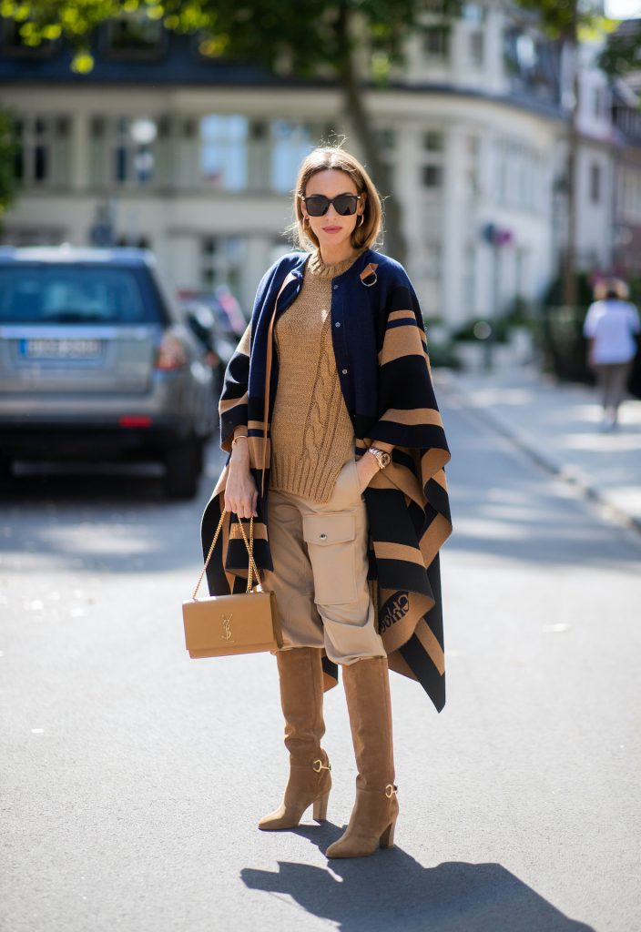 Alexandra Lapp is seen wearing Chloé striped wool cape, Chloé chunky-knit sleeveless jumper in camel, Chloé linen cargo pants in beige, SAINT LAURENT Kate medium bag in camel and CELINE Claude calfskin autumn boots in sand. All looks by Breuninger.