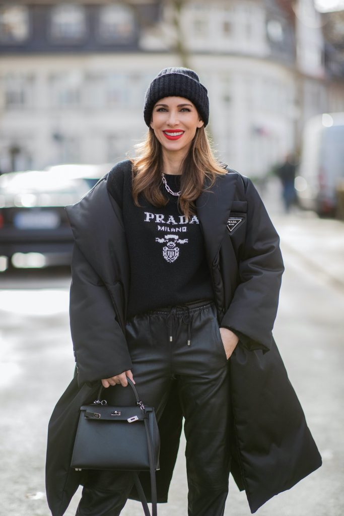 Alexandra Lapp is seen wearing one of her puffer jackets, PRADA Piumino nylon coat in black, PRADA knitted logo jumper in black, TIFFANY&CO. City HardWear necklace in sterling silver, SET leather joggers in black, XX knitted beanie, HERMÈS Kelly 28 bag in black, CHRISTIAN LOUBOUTIN So Kate pumps in black. 