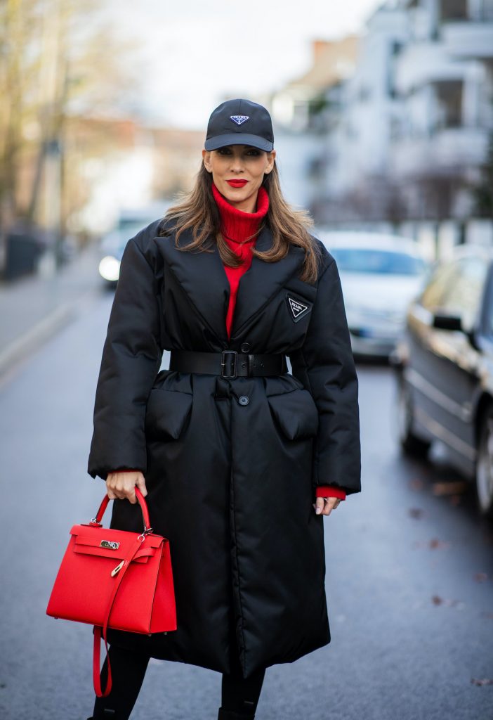 Alexandra Lapp is seen wearing one of her puffer jackets, PRADA Piumino nylon coat in black, PRADA nylon cap in black FALCONERI cashmere turtle neck dress, HERMÈS Kelly 28 bag in red, and BOTTEGA VENETA The Lug boots in black.